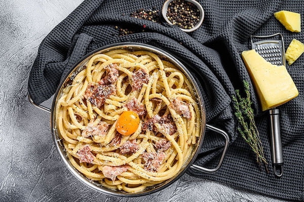 Spaghetti Carbonara mit knusprigen Speckwürfeln und glänzender Eiersauce in einer Pfanne, mit schwarzem Pfeffer
